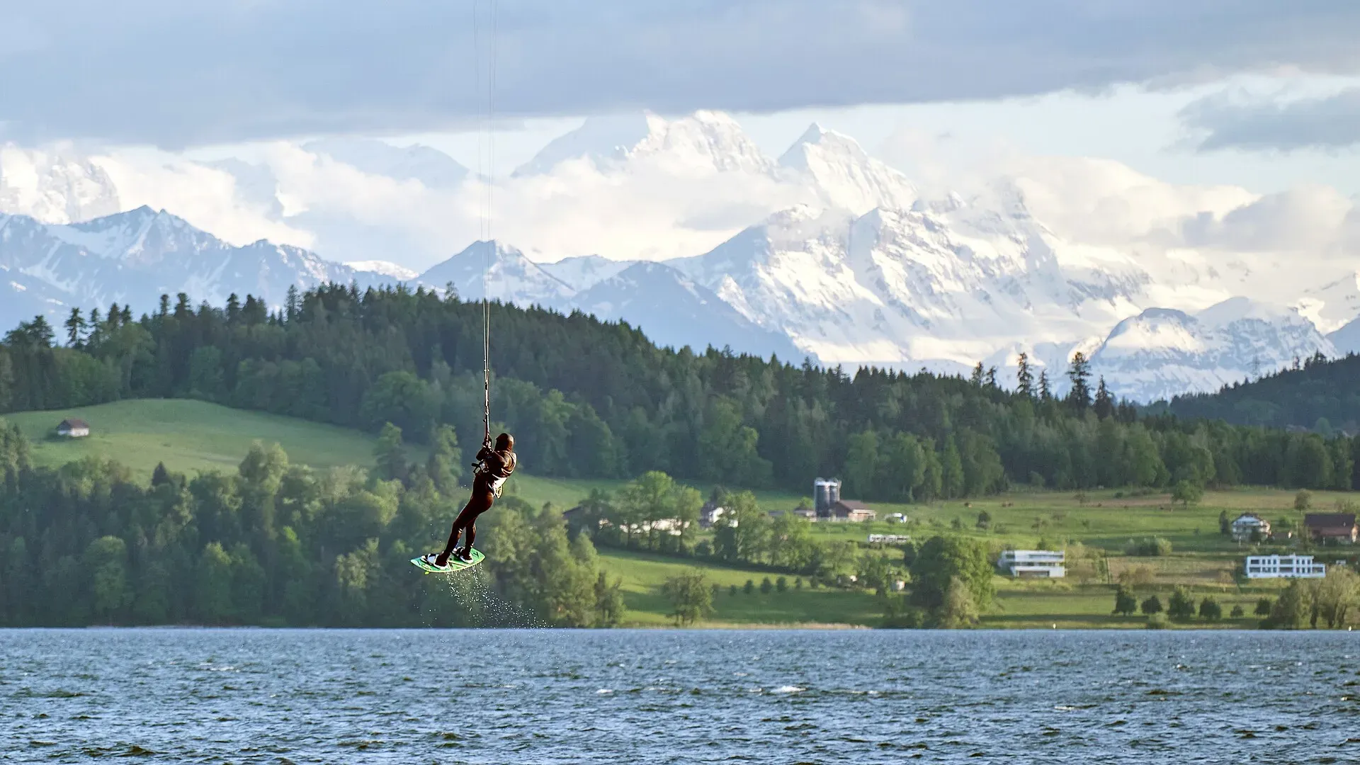 Un kitesurfeur en l’air au-dessus d’un lac suisse, forêts et Alpes enneigées en arrière-plan