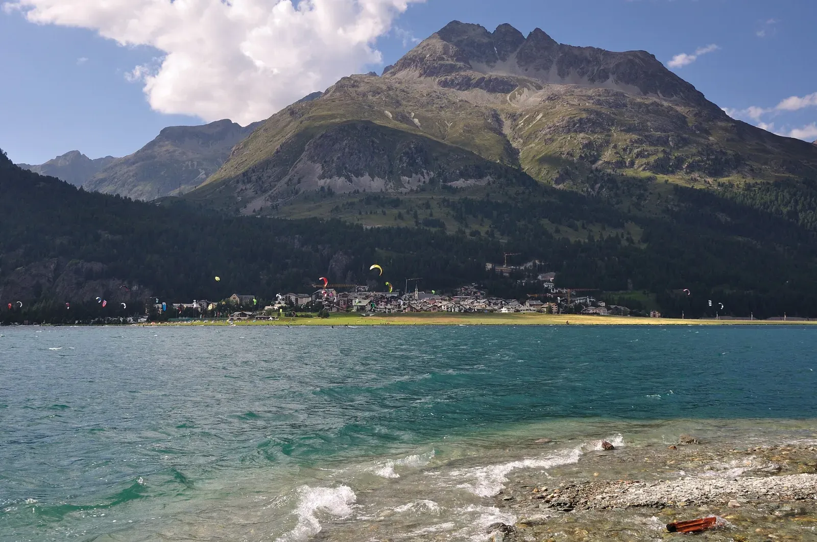 Un lac de montagne turquoise au pied d’un sommet, des ailes de kite au loin