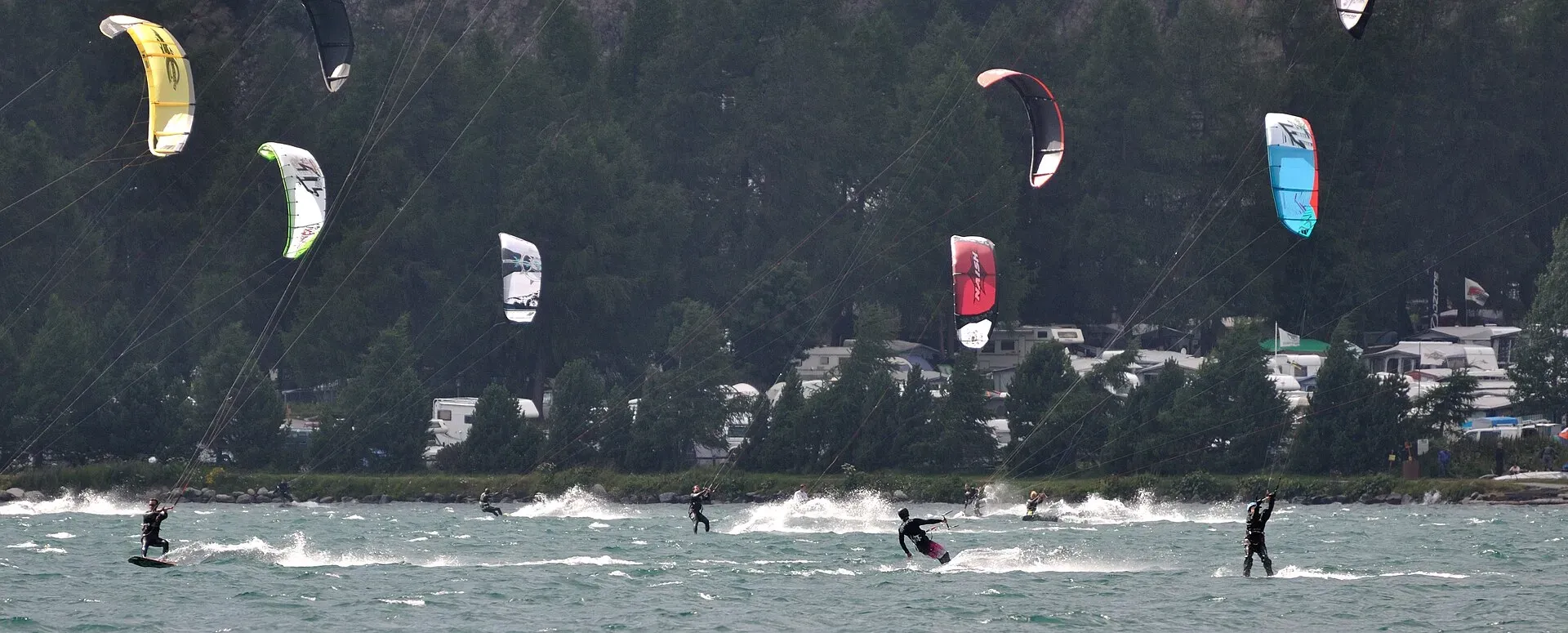 Une quinzaine d’ailes de kite colorées au-dessus d’un lac de montagne, riders en pleine vitesse