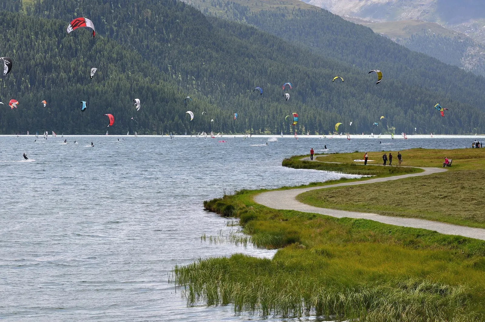 Un chemin longe la rive d’un lac de montagne où naviguent des dizaines de kites